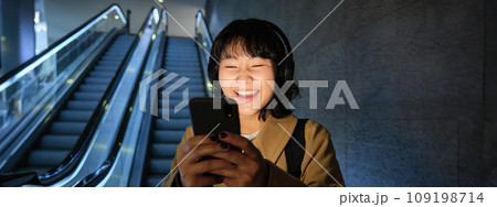 Laughing cute Korean girl on escalator, going down to tube, underground or metro, using mobile phone, watching smth funny on smartphone app Laughing cute Korean girl on escalator, going down to tube, underground or metro, using mobile phone, watching smth funny on smartphone app 109198714