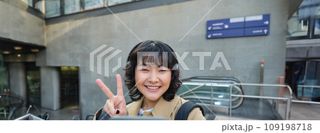 Cute and stylish Korean girl, wears headphones, takes selfie on smartphone, tourist records video or makes a photo, stands on street Cute and stylish Korean girl, wears headphones, takes selfie on smartphone, tourist records video or makes a photo, stands on street 109198718