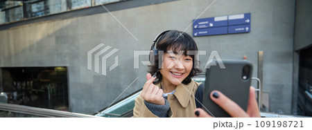 Cute and stylish Korean girl, wears headphones, takes selfie on smartphone, tourist records video or makes a photo, stands on street 109198721