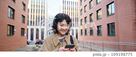Portrait of cute girl tourist, korean woman in headphones, looks at mobile phone, uses smartphone app, map or text messages, listens music in headphones Portrait of cute girl tourist, korean woman in headphones, looks at mobile phone, uses smartphone app, map or text messages, listens music in headphones 109198775