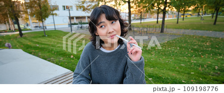 Portrait of asian woman, student in park, sits on bench with digital tablet and a pen, thinking, looking aside thoughtful, making notes, does her homework outdoors Portrait of asian woman, student in park, sits on bench with digital tablet and a pen, thinking, looking aside thoughtful, making notes, does her homework outdoors 109198776