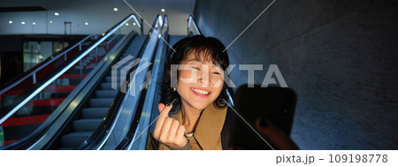 Stylish smiling asian girl, taking selfie on mobile phone while riding escalator, going down to metro station, showing finger heart and posing for photo Stylish smiling asian girl, taking selfie on mobile phone while riding escalator, going down to metro station, showing finger heart and posing for photo 109198778