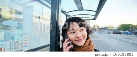 Close up portrait of cute young woman calling someone, waiting on bus stop with smartphone, using telephone while expecting public transport to arrive 109198802