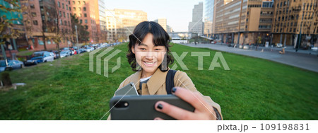 Happy young woman, student takes selfie on street, holds her tablet and homework notes, uses smartphone Happy young woman, student takes selfie on street, holds her tablet and homework notes, uses smartphone 109198831