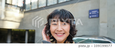 Close up of smiling brunette girl in headphones, listens music, travels around city, commutes to work, stands on a street 109199077