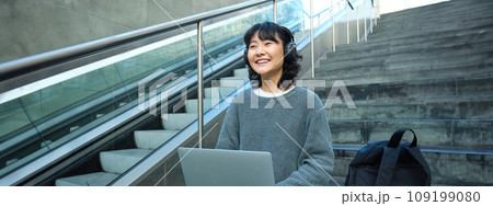Female student works on project remotely, sits on stairs street, works on laptop, listens music in headphones and smiles 109199080