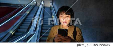 Portrait of asian girl looks shocked at mobile phone screen, standing on escalator, going down to underground or metro, commuting to work Portrait of asian girl looks shocked at mobile phone screen, standing on escalator, going down to underground or metro, commuting to work 109199085