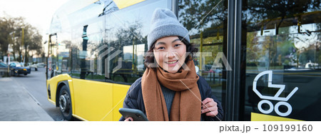 Image of girl student waiting for public transport, checks schedule on smartphone app, stands near city bus 109199160