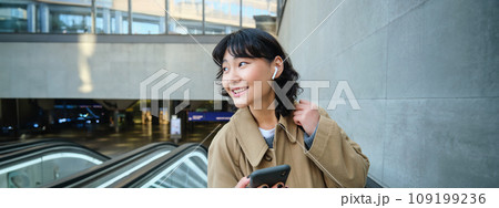 Young brunette woman commutes, goes somewhere in city, stands on escalator and uses mobile phone, holds smartphone and smiles, listens music in wireless headphones 109199236