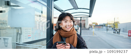 Portrait of Korean woman in winter jacket, standing with smartphone, waiting for bus on stop, looking at mobile phone app checking public transport application 109199237
