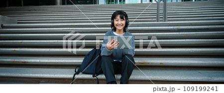 Young woman draws on her tablet, listens to music in headphones. Asian girl sits on stairs and does graphic design project, sits on street stairs Young woman draws on her tablet, listens to music in headphones. Asian girl sits on stairs and does graphic design project, sits on street stairs 109199241