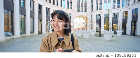 Stylish korean girl in headphones, listens music and uses mobile phone, stands in city centre, waits for someone on street and writes text message on her smartphone Stylish korean girl in headphones, listens music and uses mobile phone, stands in city centre, waits for someone on street and writes text message on her smartphone 109199253