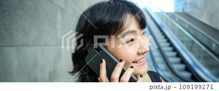 Headshot of smiling korean woman with smartphone, makes a phone call, goes down escalator in city, commutes to university Headshot of smiling korean woman with smartphone, makes a phone call, goes down escalator in city, commutes to university 109199271