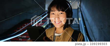 Close up of cute asian girl in headphones, picks song to listen while commuting, going down escalator to metro, smiling as reading message on mobile phone Close up of cute asian girl in headphones, picks song to listen while commuting, going down escalator to metro, smiling as reading message on mobile phone 109199303