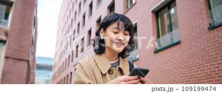 Happy korean girl walks on street, listens music in wireless earphones and holds smartphone, picks song in playlist while standing outdoor near building, reading message Happy korean girl walks on street, listens music in wireless earphones and holds smartphone, picks song in playlist while standing outdoor near building, reading message 109199474