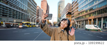 Happy asian girl tourist, takes selfie in city centre, makes video call and waves at smartphone camera, greets someone over phone 109199622