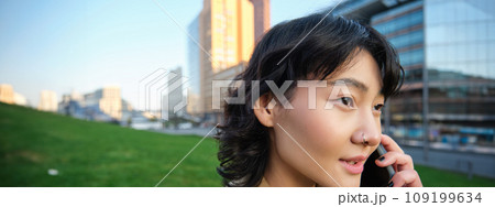 Headshot of young korean woman walks on street, talks on mobile phone, has telephone conversation, stands near campus building Headshot of young korean woman walks on street, talks on mobile phone, has telephone conversation, stands near campus building 109199634