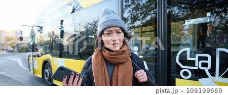 Portrait of confused asian girl, standing on bus stop, holding mobile phone, looking shocked and upset, disappointed by public transport delay 109199669