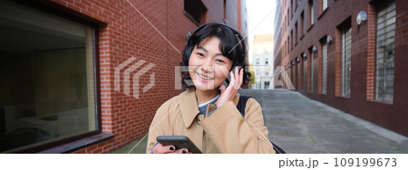 Portrait of young korean woman walking around city with backpack and headphones, listens music, looks at smartphone, uses phone application on streets Portrait of young korean woman walking around city with backpack and headphones, listens music, looks at smartphone, uses phone application on streets 109199673