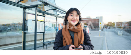 Happy smiling korean girl, using mobile phone, standing on bus stop with smartphone, looking at departure schedule on application, posing in winter clothes Happy smiling korean girl, using mobile phone, standing on bus stop with smartphone, looking at departure schedule on application, posing in winter clothes 109199830
