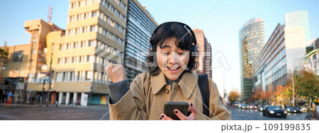 Young happy woman celebrating on street, holding smartphone and cheering, reacts amazed to good news, reads phone text message with surprised joyful face 109199835
