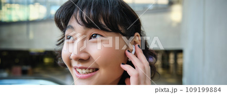 Close up portrait of smiling korean girl in headphones, listens music in wireless earphones, enjoys travelling around town with her favorite songs playlist 109199884