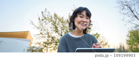 Close up portrait of young korean girl sits outdoors in park, holds her digital tablet and graphic pen, draws scatches, gets inspiration from nature for art, smiles happily Close up portrait of young korean girl sits outdoors in park, holds her digital tablet and graphic pen, draws scatches, gets inspiration from nature for art, smiles happily 109199903