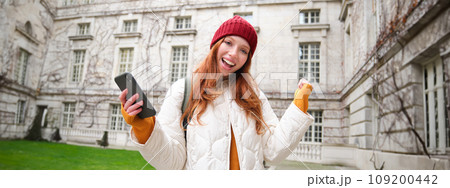 Cheerful redhead girl winning, celebrating victory, read great news on mobile phone and jumping from joy, saying yes, standing outside building in yard Cheerful redhead girl winning, celebrating victory, read great news on mobile phone and jumping from joy, saying yes, standing outside building in yard 109200442