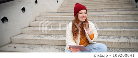Young beautiful girl with long red hair, smiles, uses internet application, connect to wifi near campus, sits on stairs with digital tablet 109200641