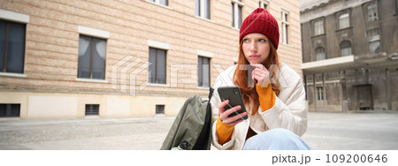 Young redhead woman with smartphone, sitting outdoors with backpack, thinking, looking thoughtful, making decision, looking at map and plans route 109200646