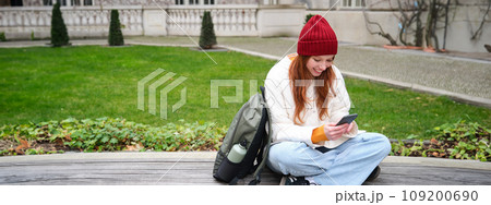 Young stylish female student with backpack, sits on bench and uses smartphone, plays on mobile phone Young stylish female student with backpack, sits on bench and uses smartphone, plays on mobile phone 109200690