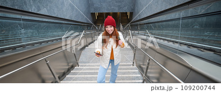 Outdoor shot of young woman plans route, follows map on smartphone app, goes up stairs with backpack and smiles 109200744