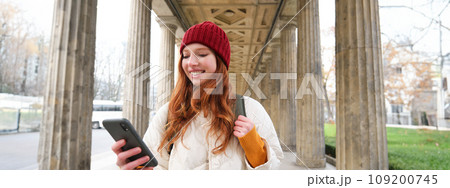 Mobile broadband and people. Smiling redhead 20s girl with backpack, uses smartphone on street, holds mobile phone and looks at application Mobile broadband and people. Smiling redhead 20s girl with backpack, uses smartphone on street, holds mobile phone and looks at application 109200745