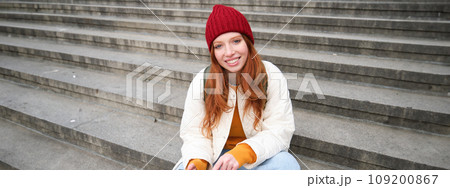 Stylish young redhead woman, talking on mobile phone app, using social media application, looking for something online on smartphone, sits on stairs outdoors Stylish young redhead woman, talking on mobile phone app, using social media application, looking for something online on smartphone, sits on stairs outdoors 109200867