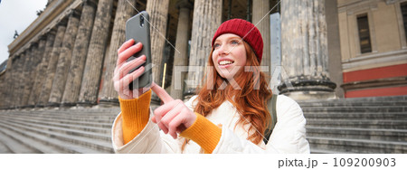 Portrait of young blogger, redhead girl tourist takes pictures of sightseeing, makes photos on smartphone camera, records video and smiles 109200903