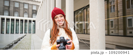 Smiling redhead girl photographer, taking pictures in city, makes photos outdoors on professional camera Smiling redhead girl photographer, taking pictures in city, makes photos outdoors on professional camera 109200941