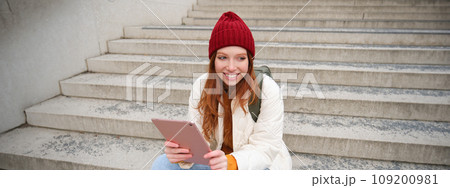 Young stylish girl, redhead female students sits on stairs outdoors with digital tablet, reads, uses social media app on gadget, plays games while waits on street Young stylish girl, redhead female students sits on stairs outdoors with digital tablet, reads, uses social media app on gadget, plays games while waits on street 109200981