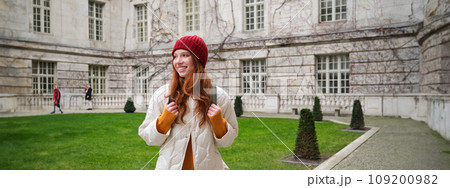 Young smiling redhead woman walking in beautiful city attractions, wearing backpack, red hat and coat, looking around with happy face Young smiling redhead woman walking in beautiful city attractions, wearing backpack, red hat and coat, looking around with happy face 109200982