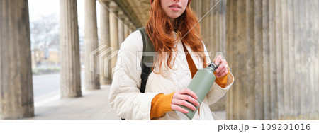 Close up portrait of redhead girl, tourist stands on street, opens thermos with hot drink, rests and pours herself coffee from flask Close up portrait of redhead girl, tourist stands on street, opens thermos with hot drink, rests and pours herself coffee from flask 109201016