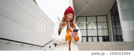 Redhead girl, young woman tourist with backpack, holds smartphone, looks for route on mobile application, searches for hotel on phone map, smiles happily Redhead girl, young woman tourist with backpack, holds smartphone, looks for route on mobile application, searches for hotel on phone map, smiles happily 109201084