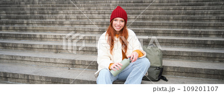 Smiling tourist, girl sits on stairs, rests on staircase, takes thermos from backpack, drinks hot coffee from flask Smiling tourist, girl sits on stairs, rests on staircase, takes thermos from backpack, drinks hot coffee from flask 109201107