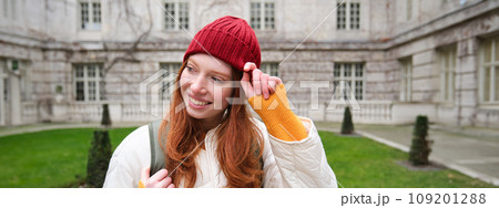 Beautiful redhead woman with backpack, tourist walking around city park and looks around, wears red hat and warm coat 109201288