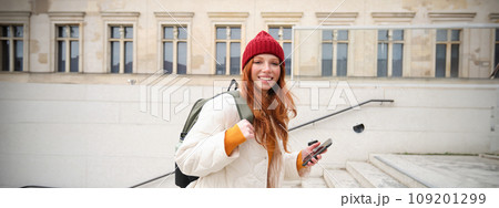 Smiling redhead girl, student tourist walks around city, goes up the stairs, looks at mobile phone map to get around town, sends message on smartphone 109201299
