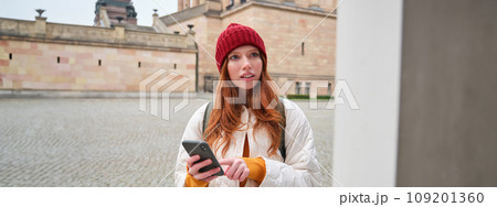 Tourism and sightseeing concept. Young redhead woman, tourist walks around city, looks at her smartphone app and at history stand, explores adventures Tourism and sightseeing concept. Young redhead woman, tourist walks around city, looks at her smartphone app and at history stand, explores adventures 109201360