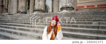 Stylish young redhead woman, talking on mobile phone app, using social media application, looking for something online on smartphone, sits on stairs outdoors 109201371
