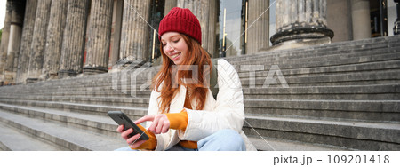 Portrait of young urban girl in red hat, sits on stairs near museum, holds mobile phone, connects to public wifi and surfs net, uses smartphone apps 109201418