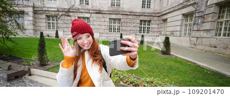 Portrait of girl blogger records video and says hi at phone camera. Young woman takes selfie with smartphone app, waves hand, video chats in application 109201470