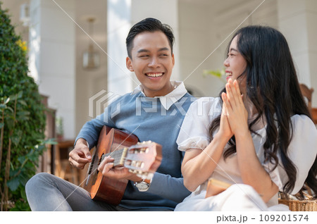 Cheerful and happy young Asian couple enjoying playing guitar and picnic at home backyard together. 109201574
