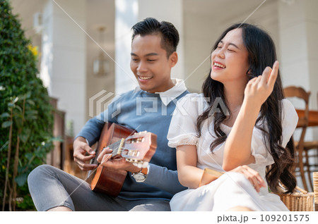 Cheerful and happy young Asian couple enjoying playing guitar and picnic at home backyard together. 109201575