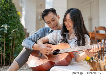 Cheerful and happy young Asian couple enjoying playing guitar and picnic at home backyard together. 109201576